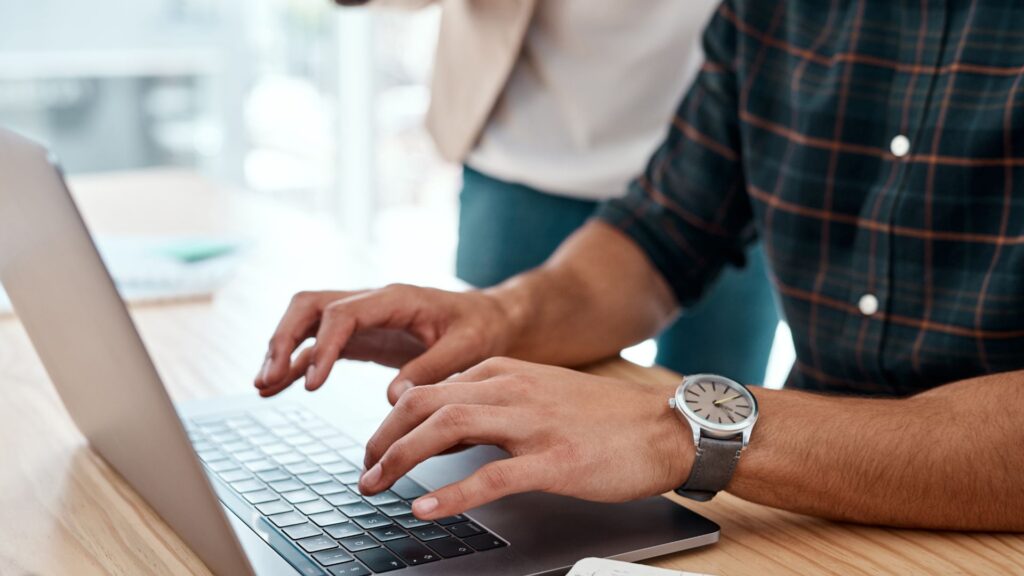 man's hands typing on a laptop keyboard while a woman stands behind him.